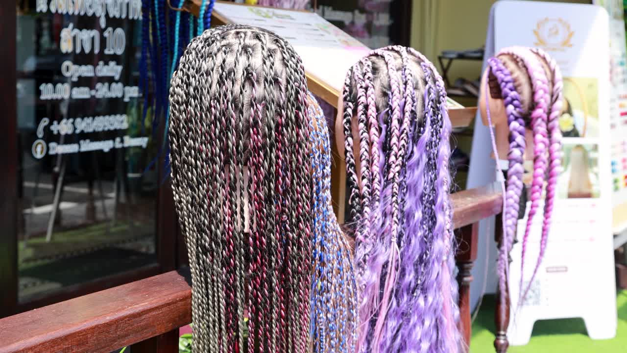 Hands interact with colorful braided hair extensions in a vibrant outdoor market setting, showcasing diverse styles and textures