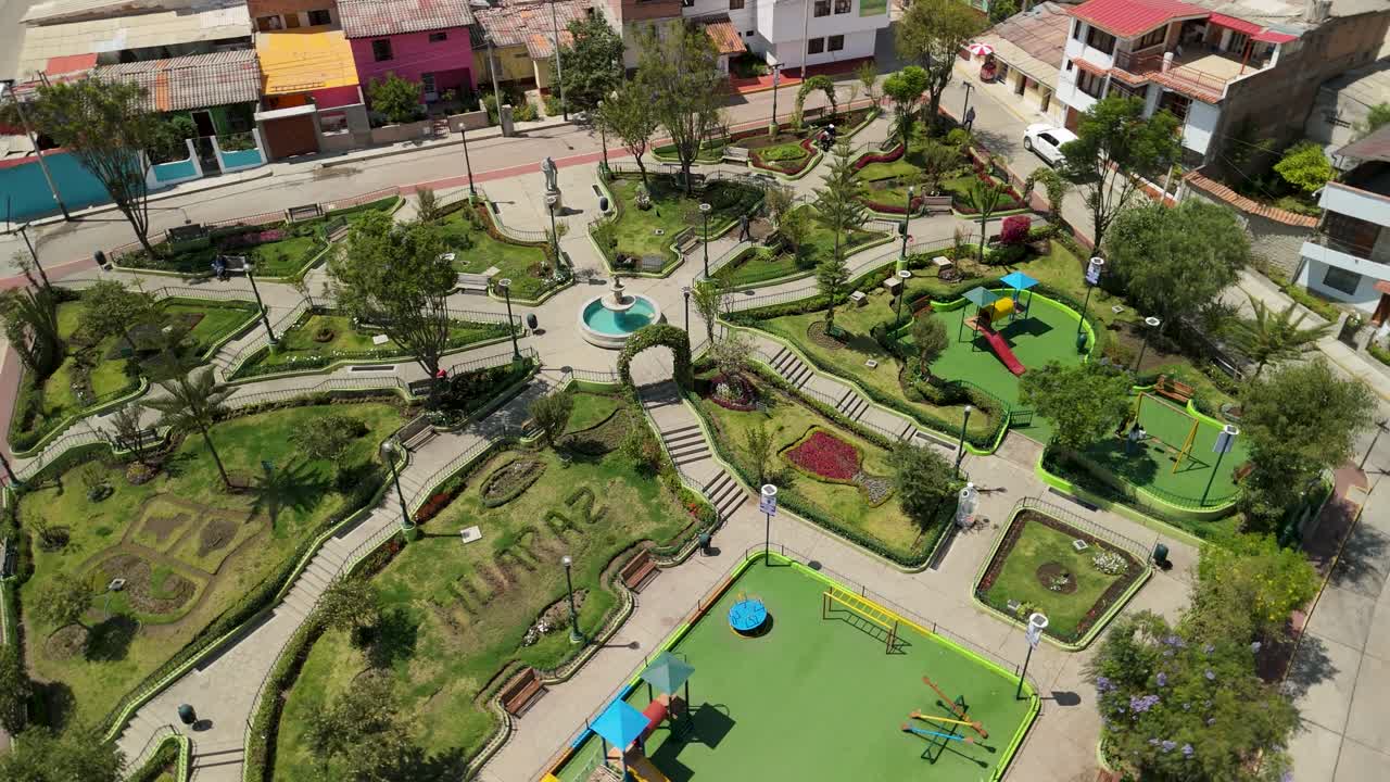 A dynamic aerial parallax shot circles over the beautifully landscaped Parque Santiago in Huaraz, Peru, revealing its intricate paths, fountain, playground, and surrounding city buildings