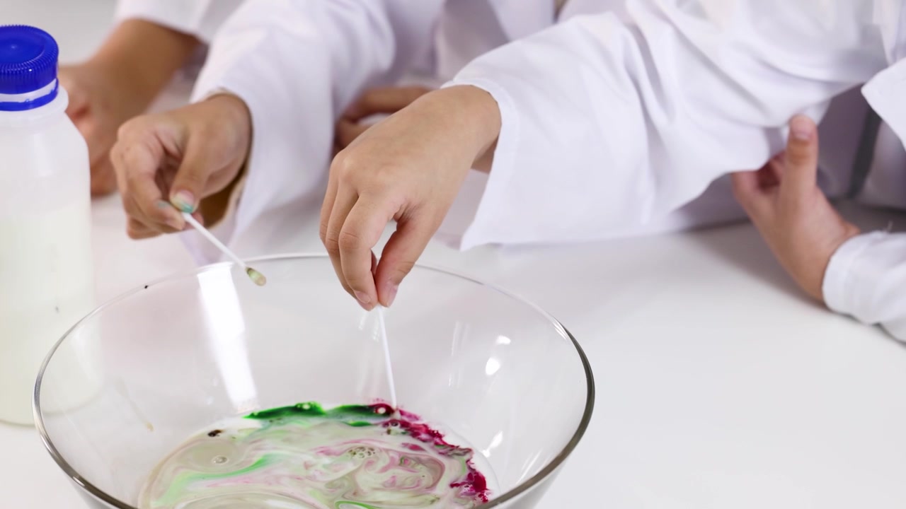 Kinder in weißen Laborkitteln rühren im Unterricht mit Wattestäbchen Lebensmittelfarbe in Milch.
