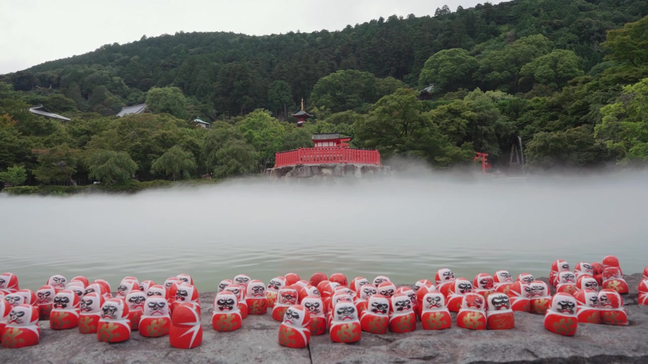 Molte bambole Daruma rosse su una piattaforma di pietra al tempio Katsuoji a Osaka, circondate da una lussureggiante vegetazione verde e una leggera nebbia, con una tradizionale struttura di tempio rosso visibile sullo sfondo