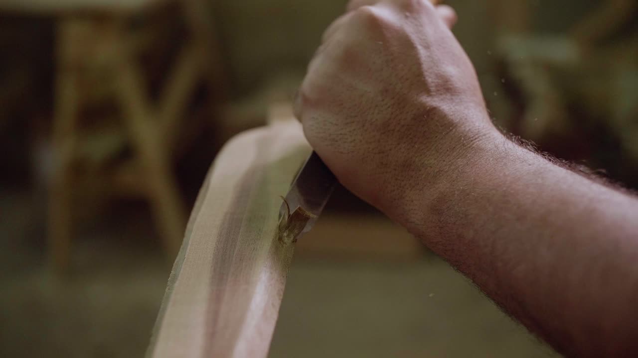 A close-up of a hand carving a piece of wood with a chisel, showing precision and craftsmanship, with wooden furniture blurred in the background.