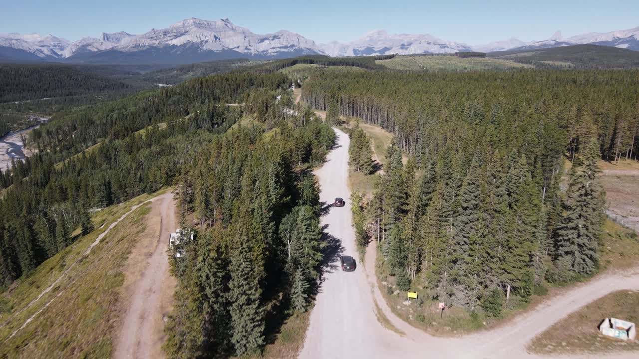dos autos conduciendo a lo largo de un polvoriento camino forestal a través de bosques de coníferas con un majestuoso telón de fondo de montañas rocosas cerca del río fantasma