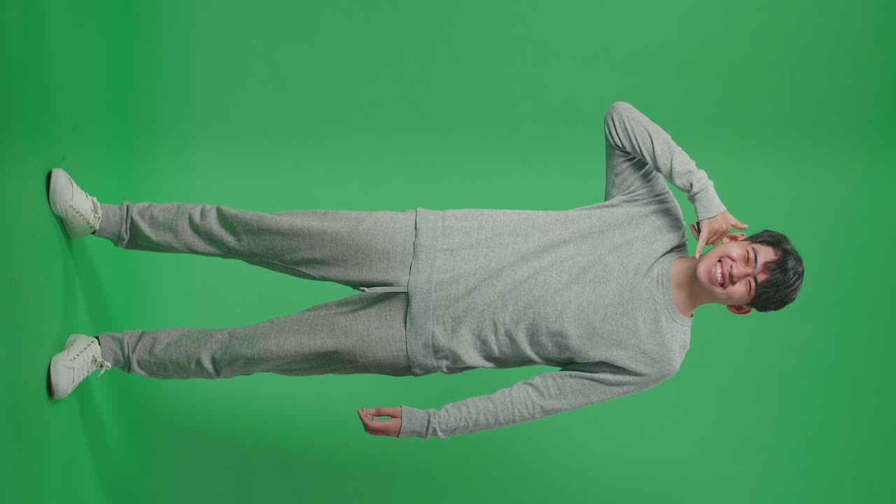 Full Body Of Cheerful Asian Man Making A Phone Call Hand Gesture While Standing In Front Of Green Screen Background