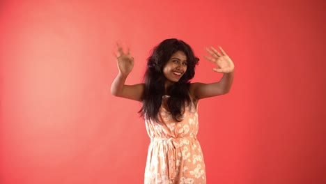 A-young-Indian-girl-in-orange-frock-saying-'Hi'-to-the-camera-standing-in-an-isolated-red-background-studio