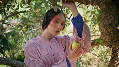 sensual fashion model standing under green tree branches holding apple close up.