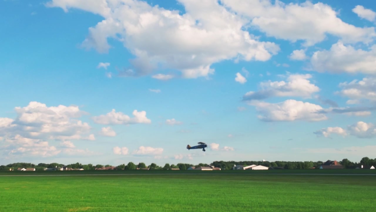 En blå biplan letter og flyver over en græsmark op i en solrig himmel.