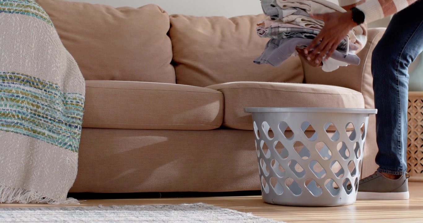 Person appearing from right is lifting folded laundry on couch and placing into basket to organize