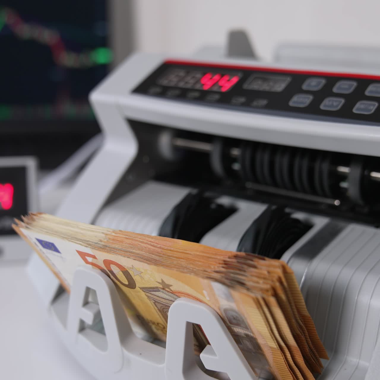Hand places a wad if fifty euro bills into a slot of cash counting machine. Counter and switched on laptop at backdrop in blur