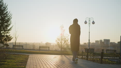 a woman walking slowly and sadly through an empty park at sunset, bathed in the warm, golden hour light. her silhouette is highlighted by the setting sun