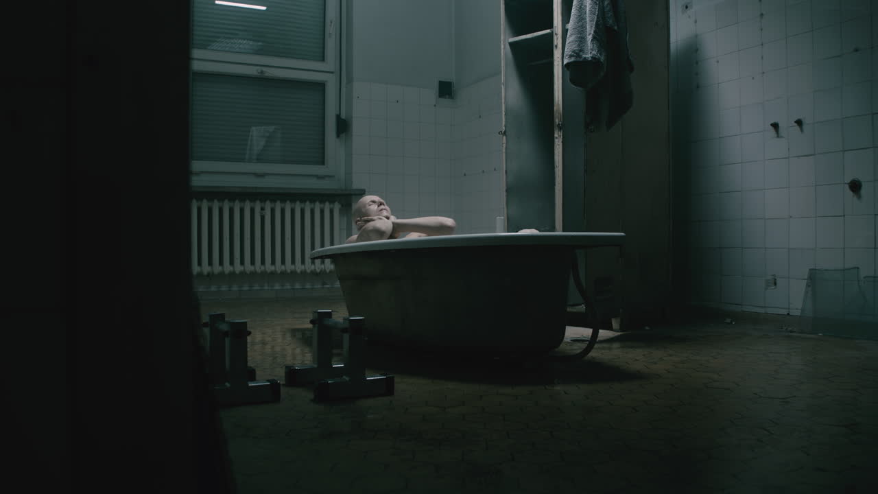 Pale bald skinny man takes a bath in the creepy old bathroom at night