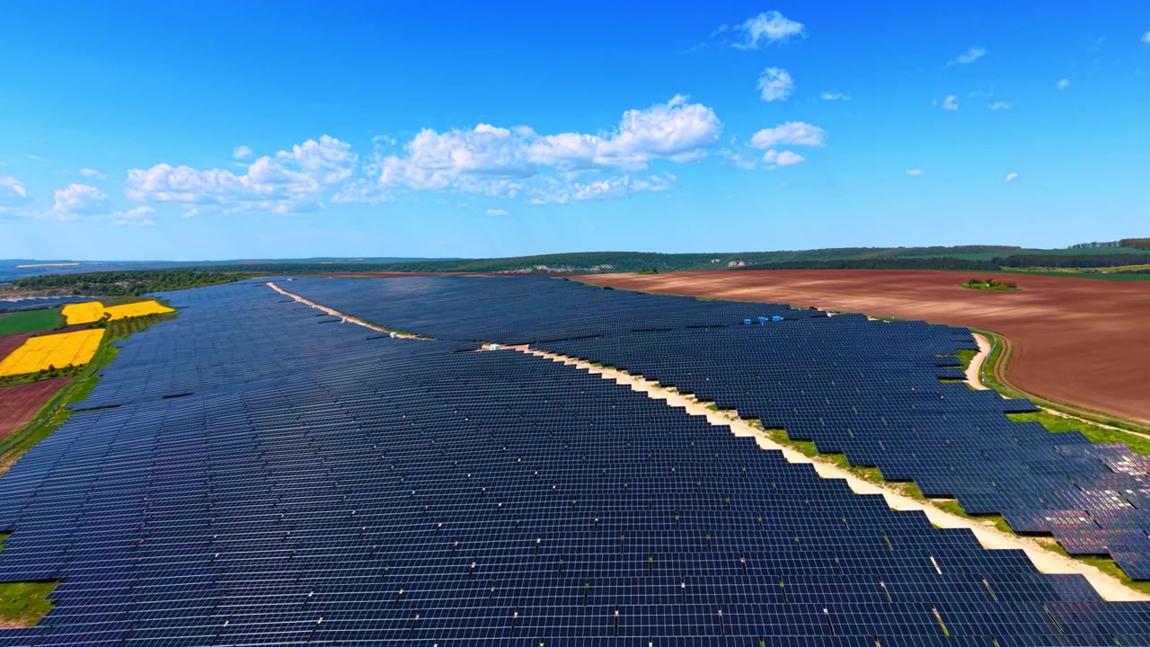 Solcelleanlæg omgivet af åbent landbrugsland, mørke rækker, jordveje og blå himmel