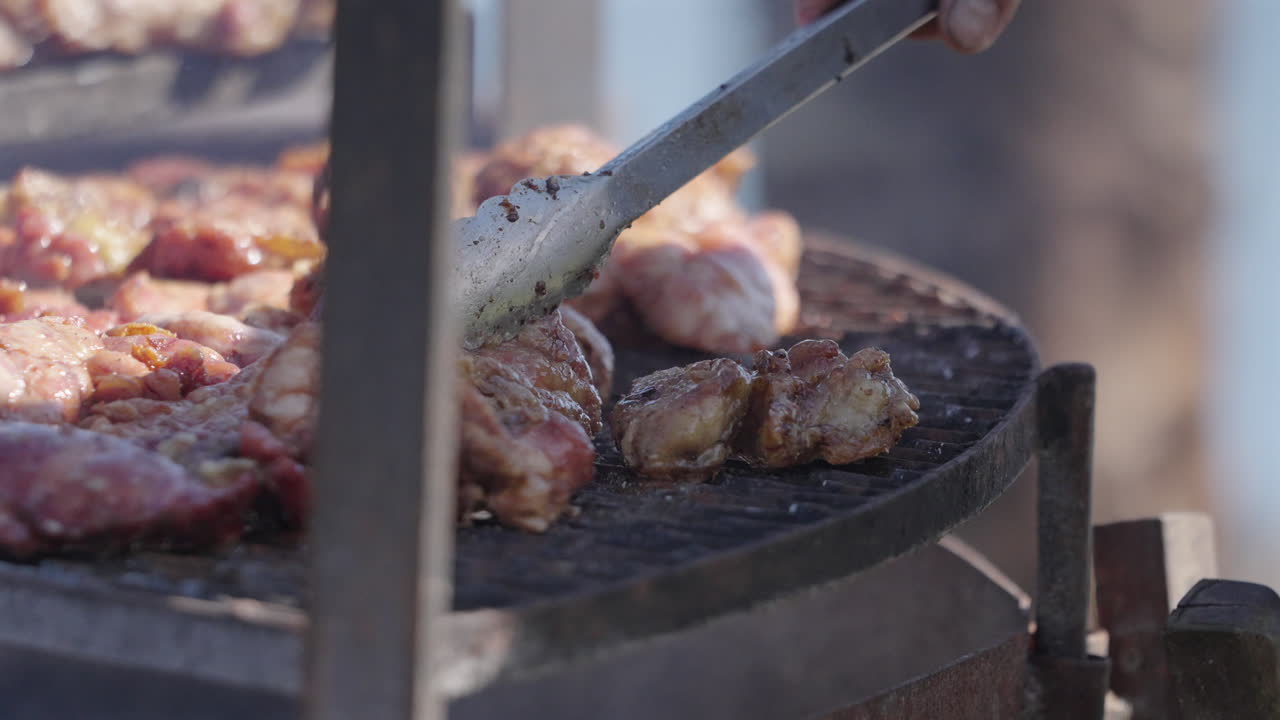Man's hand with tongs arranging grilled meat asado and chinchulines. Traditional Argentine food with burning and smoking firewood, close up