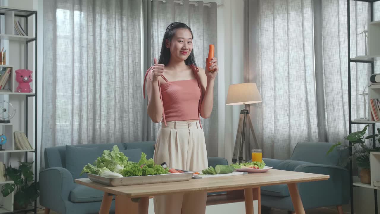 웃는 아시아 여성이 칼을 들고 당근을 잘라서 집에서 카메라에 엄지손가락을 보여주기 전에