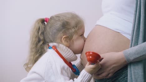 Chica-Con-Estetoscopio-De-Juguete-Besa-La-Barriga-De-Mamá-Embarazada-En-La-Habitación