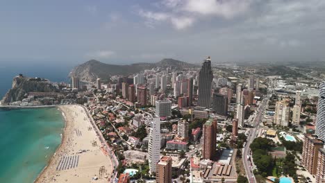 benidorm's famed coastline, spain: a high, forward drone flight offers a sweeping aerial of the beach, backed by its dense, modern skyscraper skyline on the mediterranean.