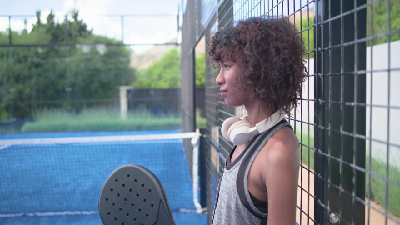 Mujer afroamericana con camiseta de tirantes apoyada en una valla golpeando una pala negra mientras la cámara se acerca, con espacio para texto.