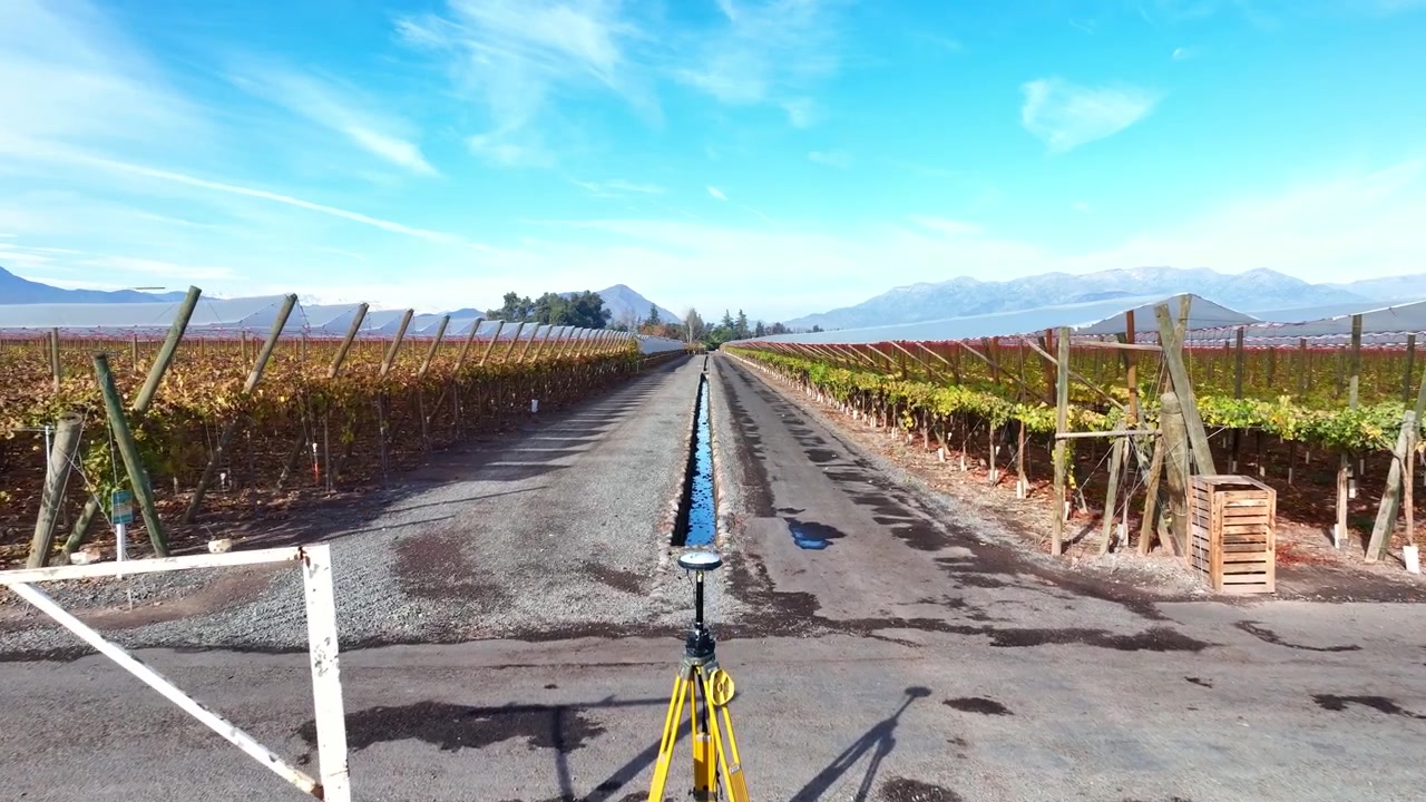 Un plano de avance lento que pasa junto a un trípode de topografía GNSS, a lo largo de un camino rural flanqueado por hileras de viñedos otoñales que convergen hacia los picos nevados de los Andes en el horizonte.