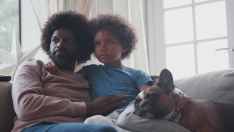 Caring-middle-aged-black-father-and-pre-teen-son-reclining-on-sofa-together-watching-TV-with-their-sleeping-pet-dog,-low-angle,-close-up