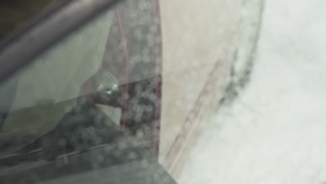 close up through car window as young adult exits red vehicle during snowfall and shuts door, water droplets scattered across glass surface, capturing cold weather