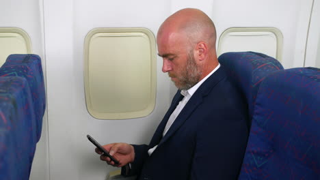 Ein-Kahlköpfiger-Mann-Mit-Bart-Und-Anzug,-Der-In-Einem-Passagierflugzeug-Ein-Mobiltelefon-Benutzt