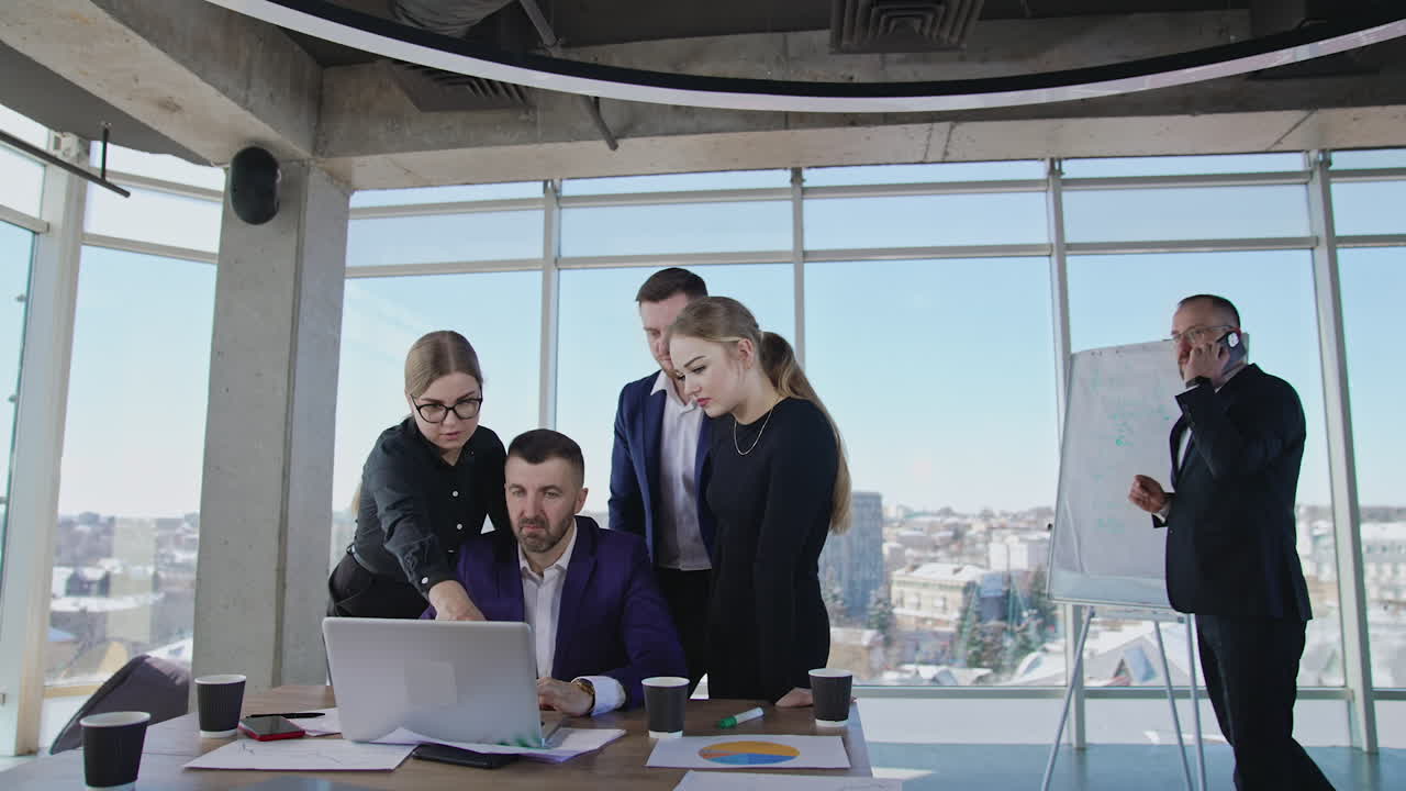 Office staff looking for new ideas and brainstorming. Male boss sitting in front of laptop and employees standing around him and making suggestions. Man speaking on the phone at the backdrop.