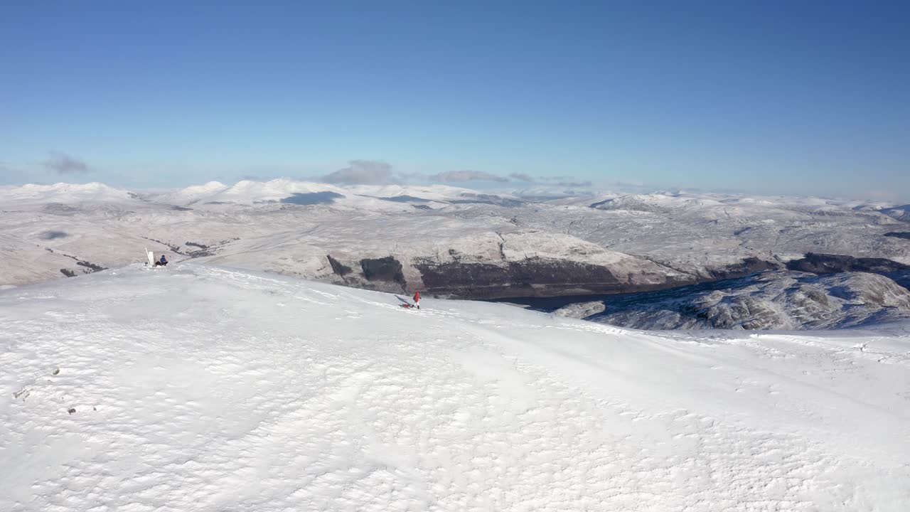 Person går tur med hund på snødekt fjelltopp under blå himmel