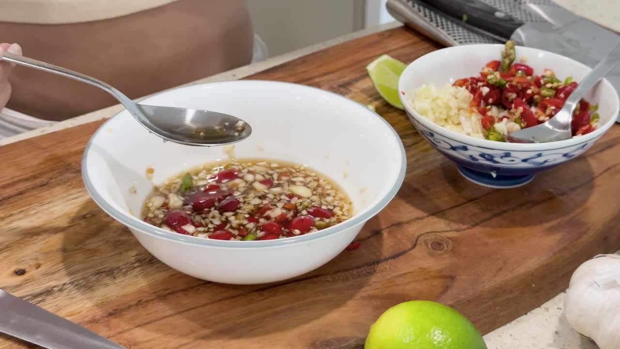 A person uses a spoon to taste freshly prepared chili fish sauce on a wooden countertop, with natural lighting and close-up camera framing