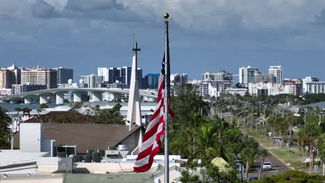 waving american flag in sarasota city during cloudy day. traffic on street of st. armands district and skyline of downtown in background. patriotic country of usa. aerial close up shot.