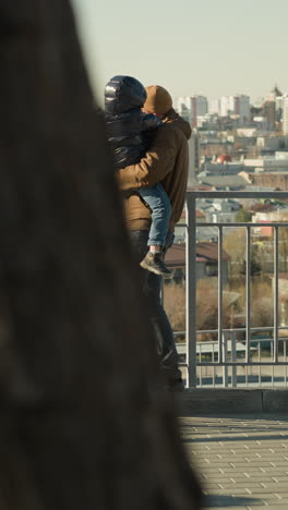 a father holds his young son close to an iron railing as they both gaze out over a cityscape, both are dressed in warm jackets, with view of cars passing by