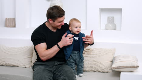 man in black t-shirt sits on the sofa holding his son by the hands. cute infant learning to stand.