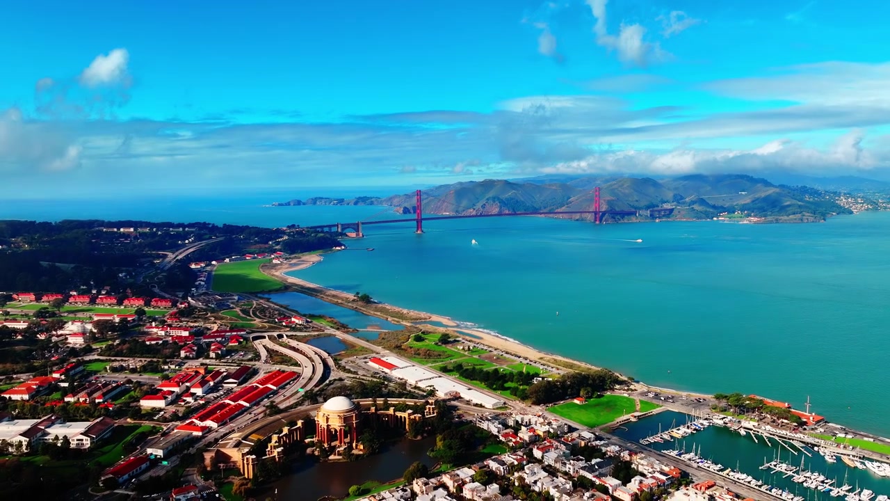 San Francisco, Kaliforniya'da Golden Gate Köprüsü ve Presidio kıyı şeridinin havadan görünümü. Mavi gökyüzü altında kırmızı çatılı kale binaları, kumlu koy, turkuaz körfez ve Marin tepeleri üzerinde drone perspektifi.
