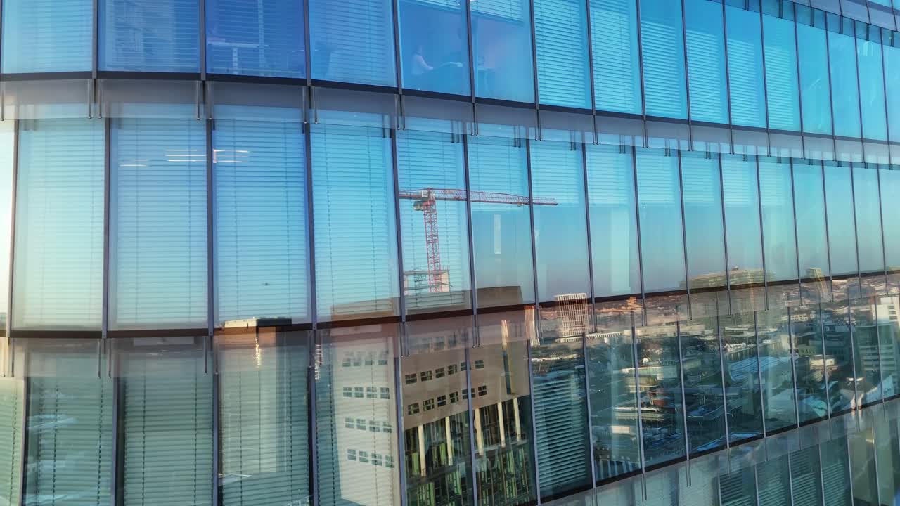 Drone inclina-se para cima ao longo de vidro refletivo, revelando interiores de escritórios e pequenos trabalhadores atrás das janelas.