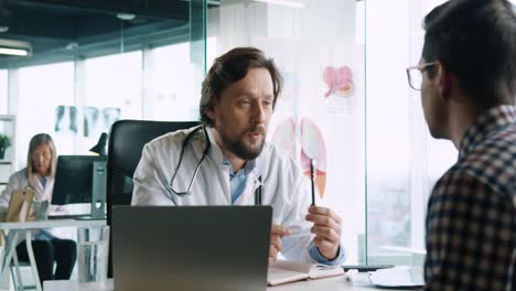 Close-up-view-of-caucasian-male-doctor-sitting-at-desk-with-laptop-and-explaining-to-male-patient-treatment-for-coronavirus-in-medical-consultation
