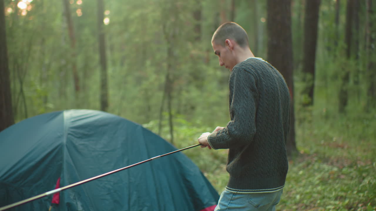 Tourists working together to fix flexible tent pole while walking toward pitched tent in forest, surrounded by trees and greenery, collaborating calmly during outdoor camping setup