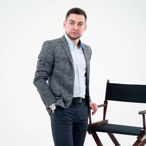 portrait of a young businessman in light studio. handsome man in elegant costume standing near the chair and looking at camera.