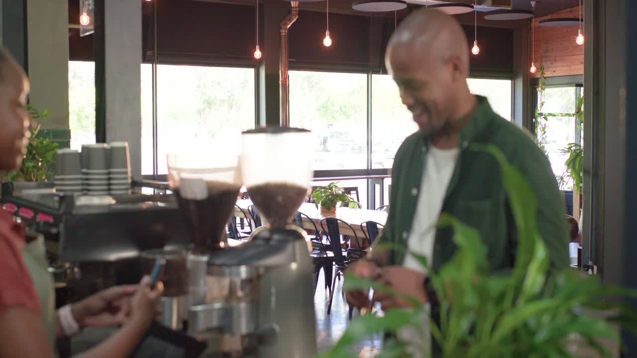Barista afroamericano in grembiule che accoglie il cliente al bancone, e il cliente che paga con la carta contactless
