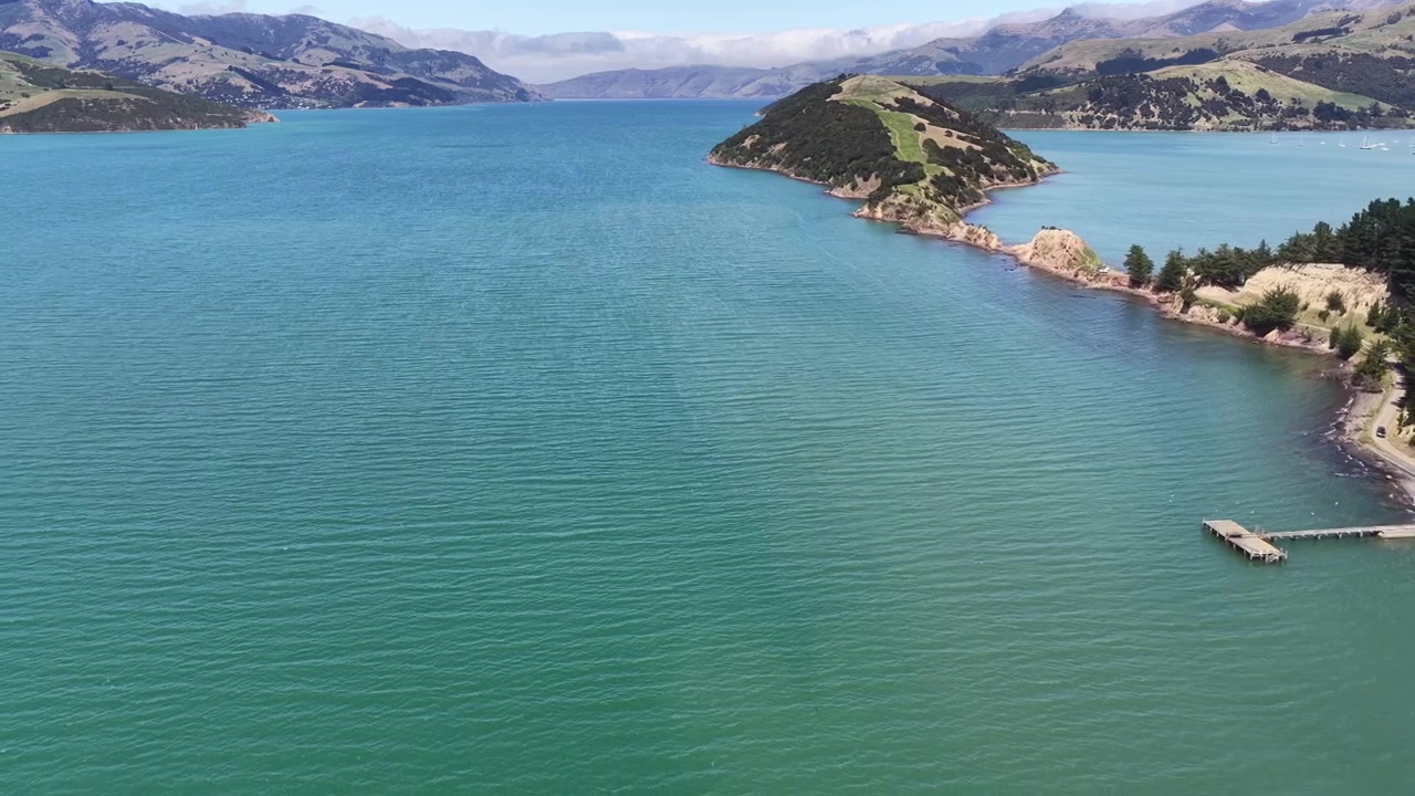 Volo aereo sopra una stretta penisola che si estende in acque blu vivide sull'Isola del Sud della Nuova Zelanda, catturando la costa frastagliata e il pacifico paesaggio costiero.