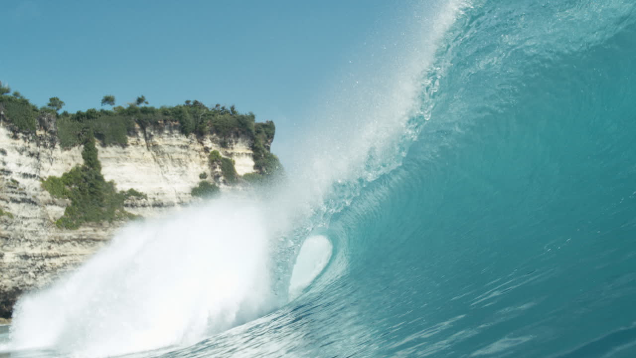 Turquoise wave curls in slow motion beneath clear blue sky and coastal cliffs