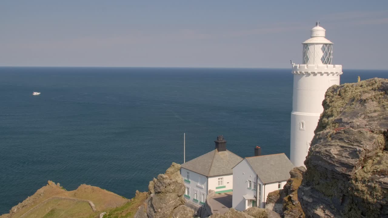 Locked-off wide shot of white lighthouse by sea, clear skies and rocky coastline