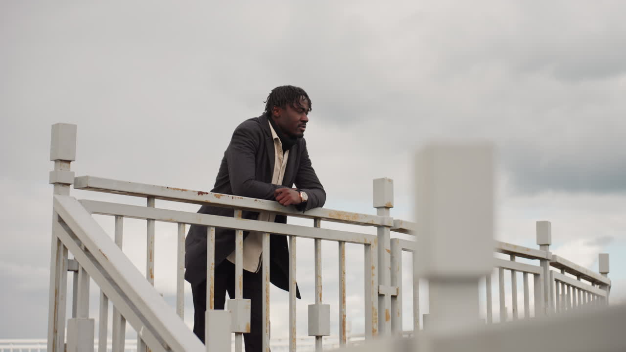 Dark skinned man stands by metal railing lost in deep thoughts as gentle breeze sways jacket, creating mood of reflection and calmness under cloudy sky while soft light enhances emotional atmosphere