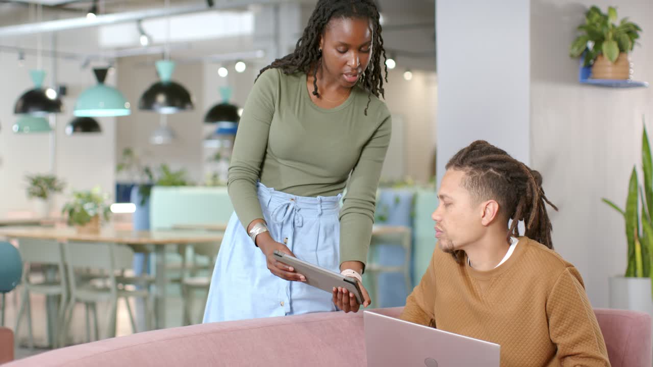 Membro del team afroamericana che mostra un tablet mentre un uomo guarda il laptop, esaminando il progetto nel salotto