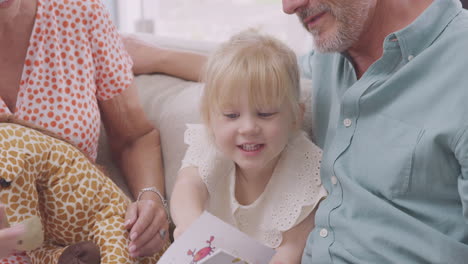 close up of grandparents sitting on sofa with granddaughter at home reading book together