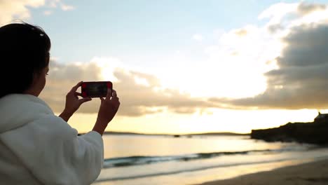 brunette woman taking a photograph of the sunset on her smartphone