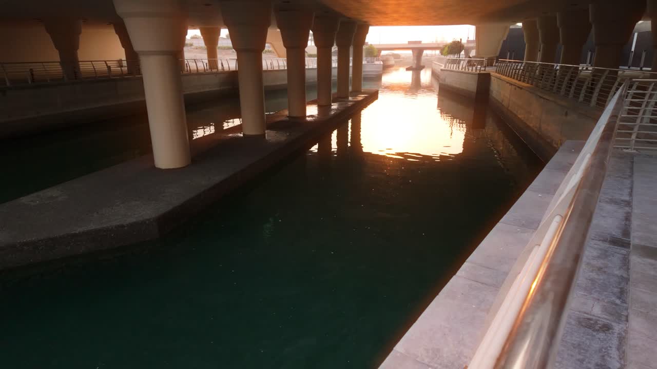 Stunning golden sunrise illuminates a peaceful sea canal under a highway bridge. The warm light reflects on tranquil waters, creating a serene, atmospheric scene