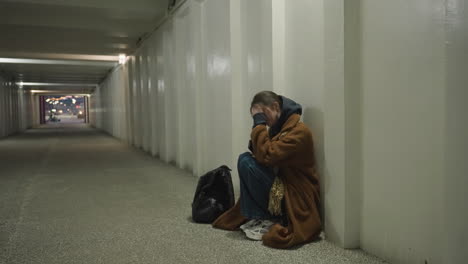 a girl in a brown coat sits bent over in an underpass tunnel, her hand covering her face while holding her phone. her expression conveys deep disconsolation and trouble