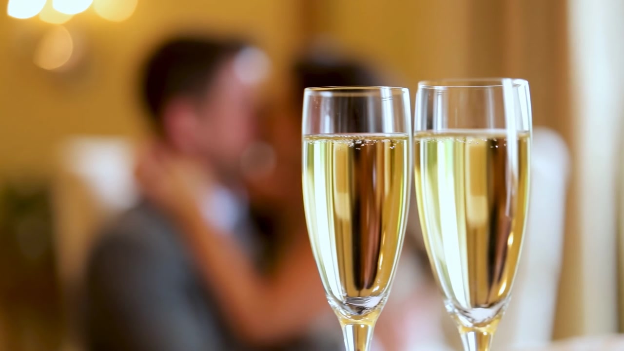 Coppia di sposi romantica che si scambia un bacio gioioso e un abbraccio in una sala banchetti, circondata da calici di champagne e sedie da pranzo, che cattura un momento celebrativo perfetto in abito bianco.