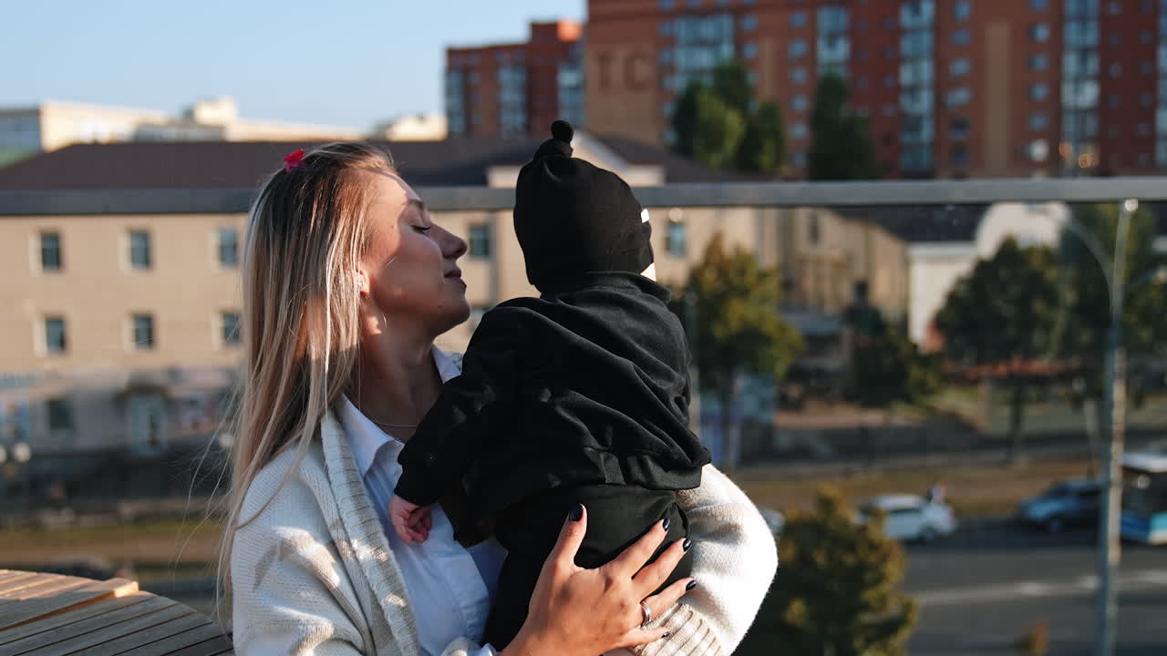 Young Caucasian woman wearing white sweater holds her baby in black clothes. Mother talks to her infant standing outdoors in the city.