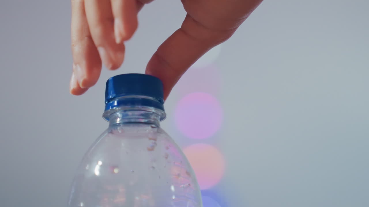Lähikuva valkoisesta kädestä sulkemassa muovipulloa sinisellä korkilla, taustalla värikkäitä bokeh-valoja, vesipisaroita pullon sisällä, hellävarainen kiertoliike, ASMR-tyydyttävä tunnelma, nopea kuvasarja.
