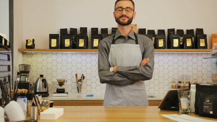 Giovane barista caucasico con gli occhiali in posa per la fotocamera con le braccia incrociate dietro il bancone di una caffetteria
