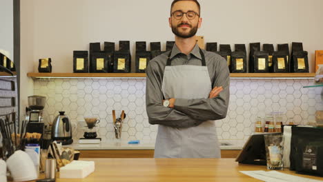 young caucasian male barista wearing glasses posing for the camera and crossing arms behind the bar of a coffee shop